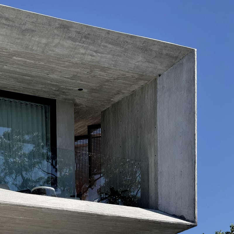 Corner of a board-formed concrete upper level with a deep overhang, glass balustrade and a timber lattice screen against a clear blue sky