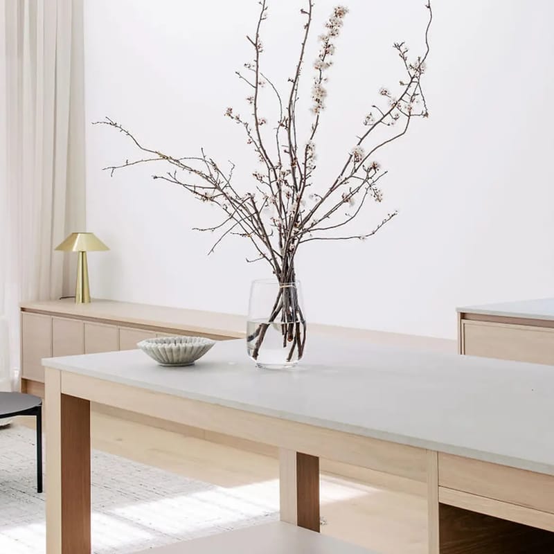 Light-filled kitchen and living area with a pale stone bench, timber joinery and a vase of blossom branches