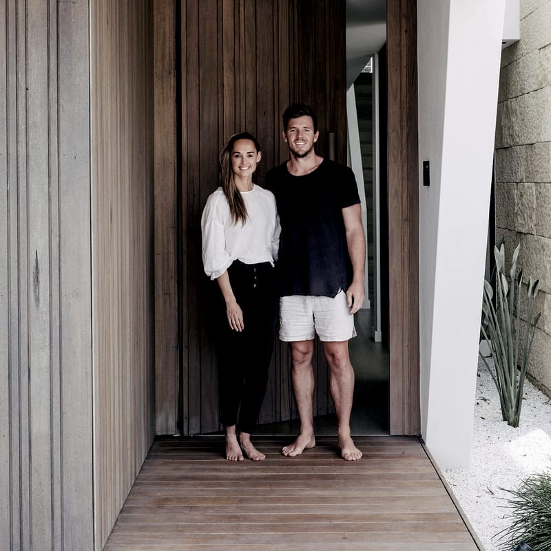 A couple standing barefoot in a timber-lined entry beside a tall timber pivot door, white render and a stone wall with coastal planting